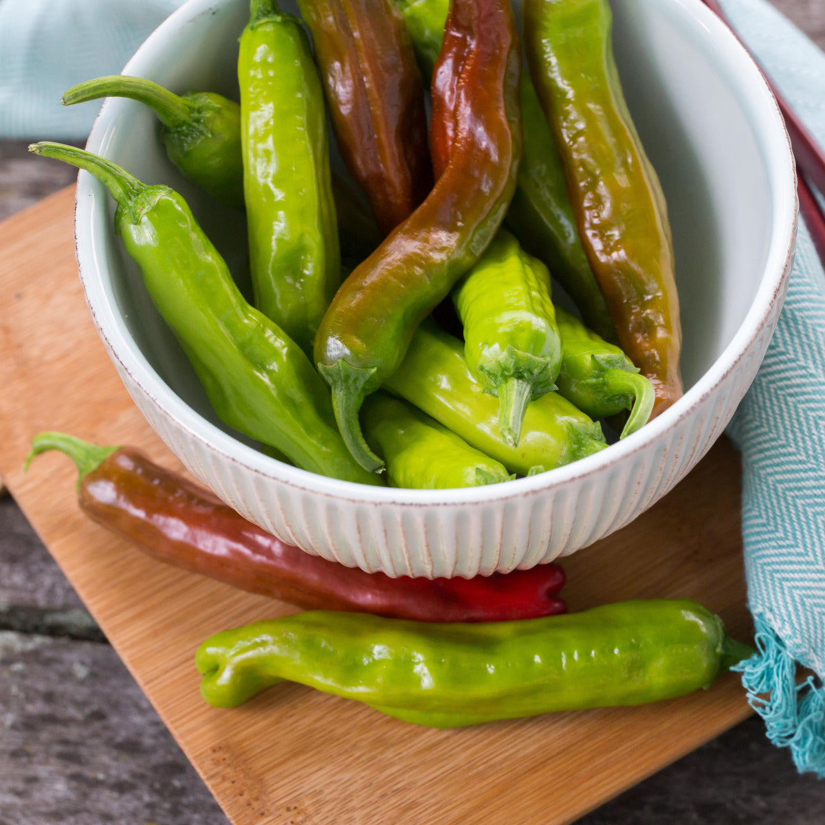 A white bowl filled with green and partially red Dragon Roll' Peppers (Capsicum) sits on a wooden surface with extra peppers beside the bowl - Photo Courtesy of Ball Horticulture, Inc. Decorative pot not included.
