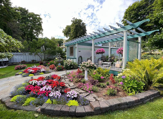 A vibrant garden with colorful flowers and green plants surrounds a patio covered by a pergola. The area includes outdoor furniture, hanging flower baskets, and a white picket fence in the background.