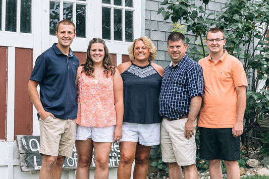 Five people stand close together and smile in front of a house with white and red doors. They are dressed in summer clothes, and there are green plants and a rustic wood sign in the background.