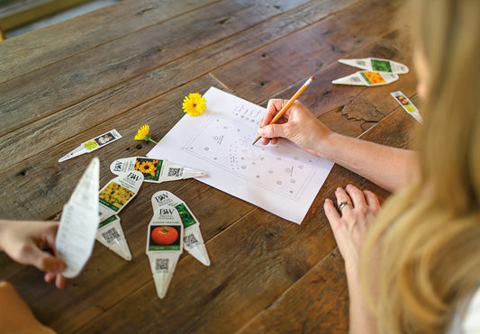 Two people sit at a wooden table, planning a garden on paper with plant markers and seed packets spread around, while one person writes and a yellow dandelion lies on the paper.