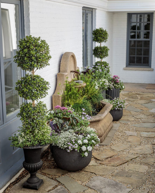 A stone fountain with water flowing into a trough is surrounded by potted topiary trees and flowering plants on a stone patio beside a white brick house with blue-gray window frames.