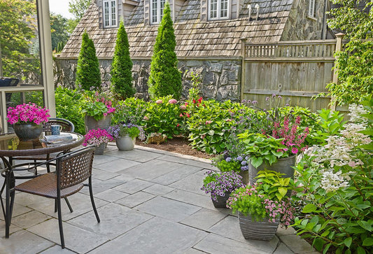 A stone patio with a table and chairs is surrounded by potted and garden flowers and greenery. A wooden fence and stone building are in the background, with tall evergreens and blooming plants adding color and texture.