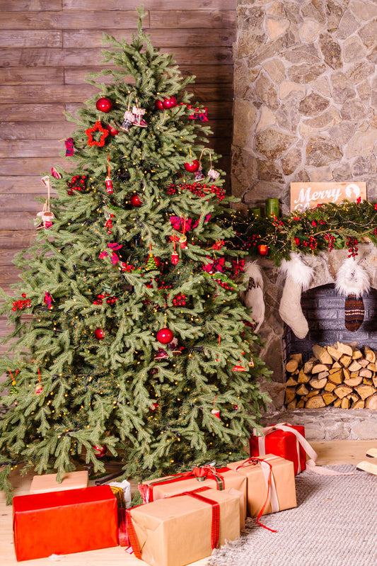 A decorated Christmas tree with red ornaments and gold garlands stands beside a fireplace. Wrapped gifts in brown and red paper are placed underneath the tree. The mantel is decorated with stockings and festive decor.
