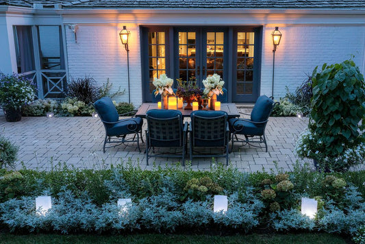 A cozy outdoor patio with four cushioned chairs around a table, decorated with flowers and lanterns, sits on a stone terrace beside a house with French doors, illuminated by wall lights and surrounded by lush landscaping.