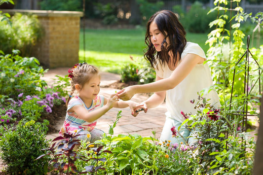A woman and a young girl are gardening together outdoors, surrounded by green plants and flowers. The woman is handing something small to the girl, who looks focused and engaged. Sunlight brightens the garden scene.