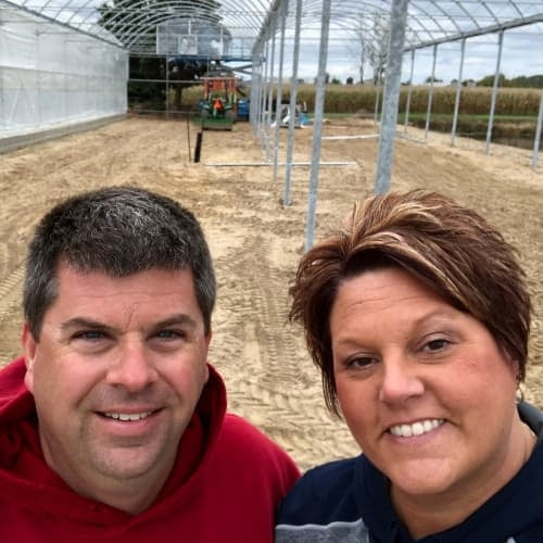 A man and a woman smile for a selfie inside a partially constructed greenhouse on a dirt lot, with metal framework and farm equipment visible in the background.