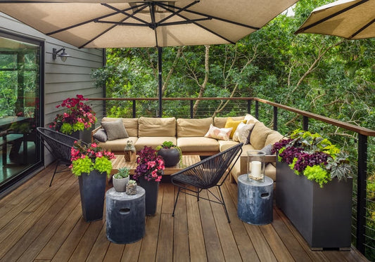 A cozy outdoor patio with a beige sectional sofa, potted plants, black chairs, round side tables, and large umbrellas, set on a wooden deck surrounded by lush green trees.