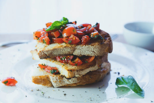 Three slices of toasted bread stacked on a plate, topped with chopped tomatoes, basil, and herbs, resembling bruschetta. A basil leaf and tomato pieces are on the plate, with soft lighting in the background.