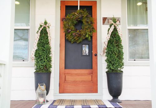 A front door decorated with a large holiday wreath. Two tall potted evergreen shrubs with ribbon bows flank the door, and a lantern sits on the porch beside a striped welcome mat.