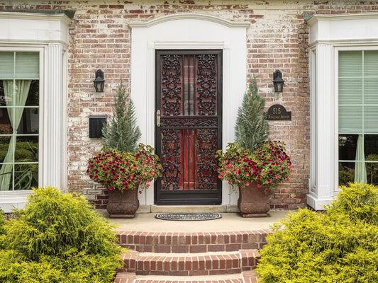 A brick house entrance with a black ornate metal door, two large planters with red flowers and greenery on either side, and white columns framing the doorway. Steps and neatly trimmed bushes lead up to the door.
