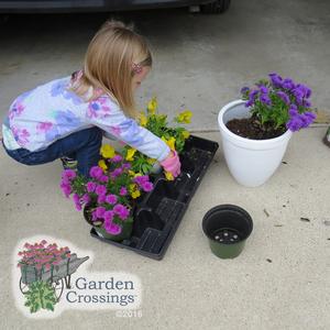 A young girl with blonde hair bends over a white pot of yellow and purple flowers on a driveway, with a bag of potting mix and garden supplies nearby.