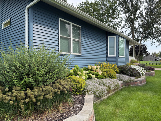 A blue house with white trim is bordered by a well-maintained garden filled with green shrubs, flowering plants, and a curved stone edge, set in a lush, grassy yard.