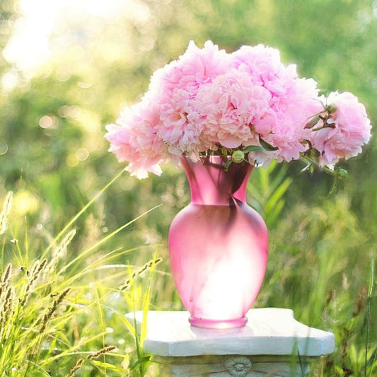 A pink frosted glass vase filled with large pink peonies sits on a small pedestal outdoors, surrounded by green grass and bathed in soft sunlight.