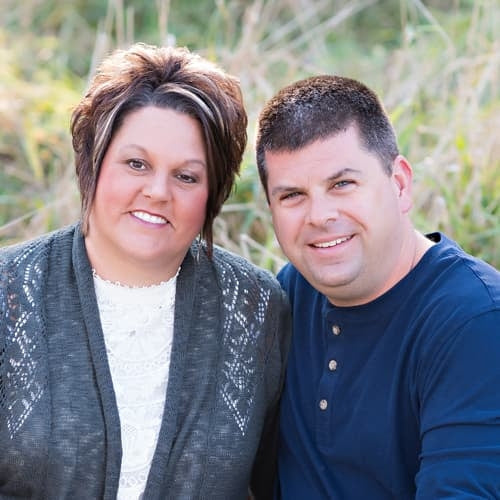 A smiling man with short hair in a blue shirt stands next to a smiling woman with shoulder length brown hair in a white shirt and grey sweater