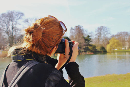 Photo from behind of a blonde woman with her hair tied up in a bun snapping a photo of a lake-side nature scene.
