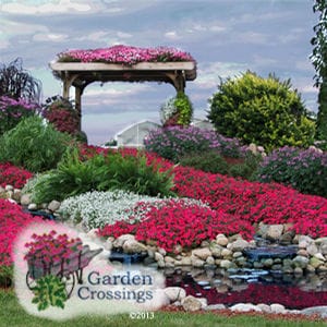 Hillside garden filled with pink and white supertunias and other green plants. A small pond is in the foreground, and a gazebo covered in flowers sits at the top against a cloudy blue sky.