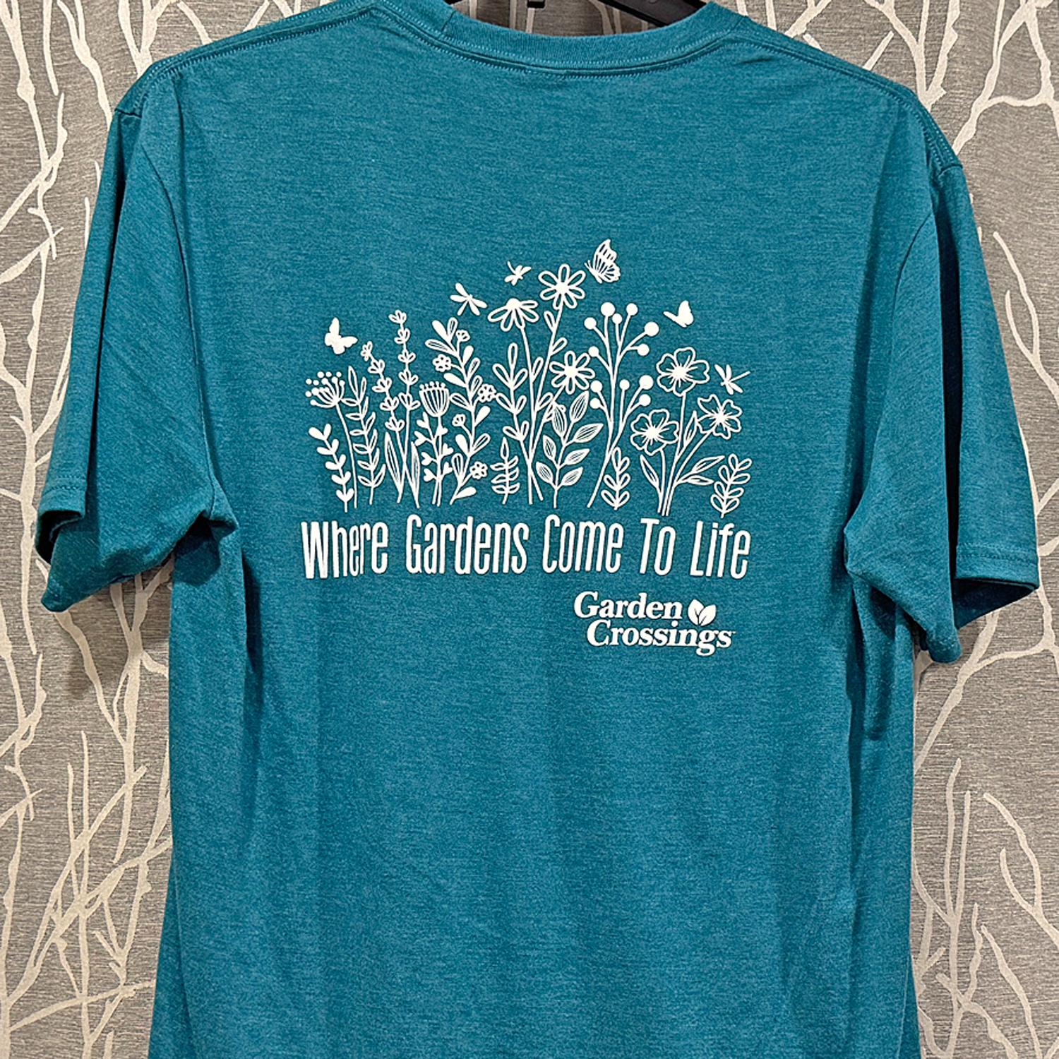 Teal t-shirt with white illustrations of flowers, butterflies, and bees. Text reads, Where Gardens Come To Life and Garden Crossings below the design. Shirt hangs on a hook against a patterned background.