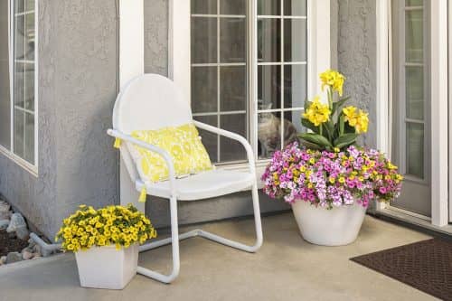porch with a white chair, white container of yellow petunias on one side and a white container of pink petunias on the other