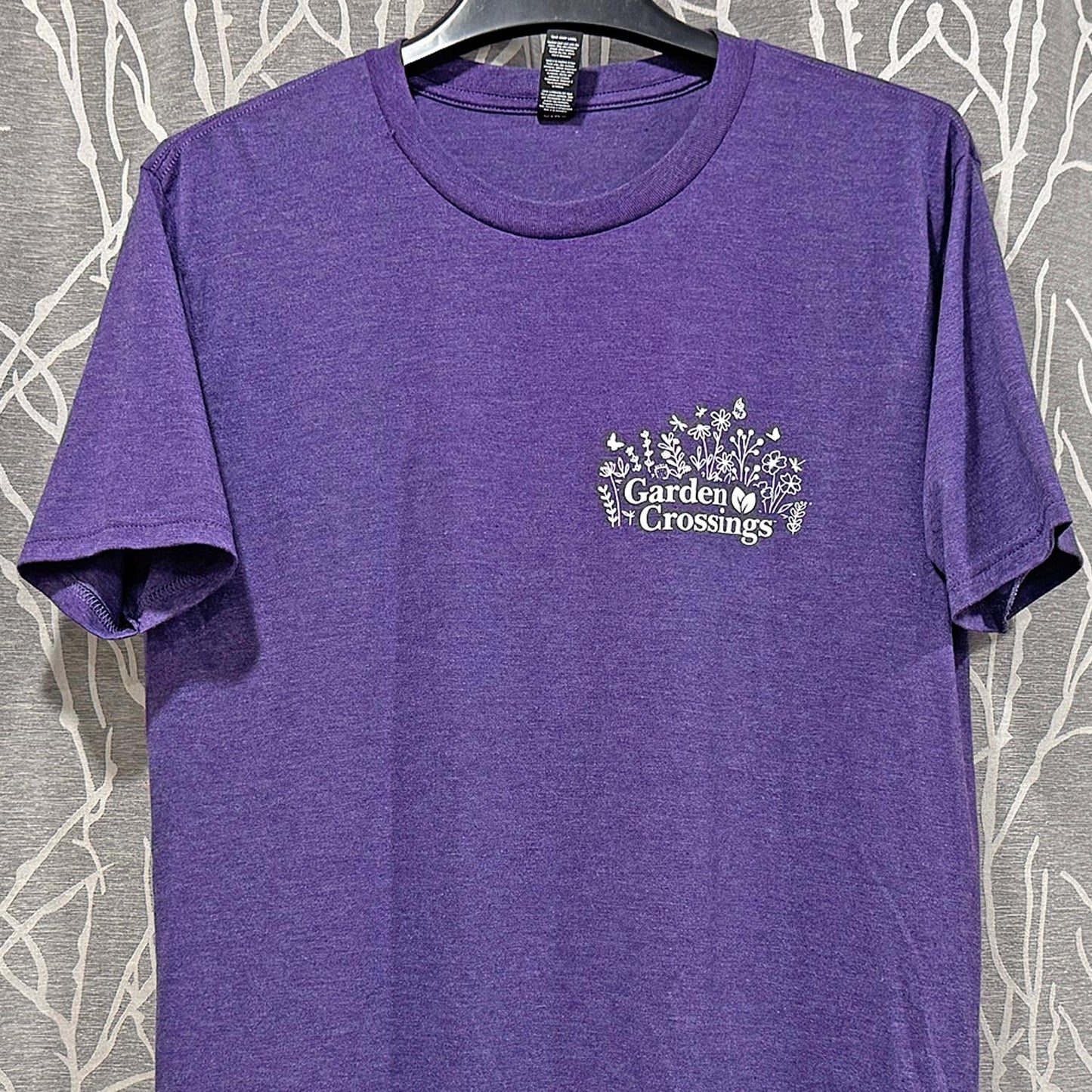 The Garden Crossings Tshirt is a purple, soft tri-blend tee with a white floral graphic and "Garden Crossings" on the left chest, shown on a hanger before a white branch pattern—ideal for gardening t-shirt enthusiasts. - 
