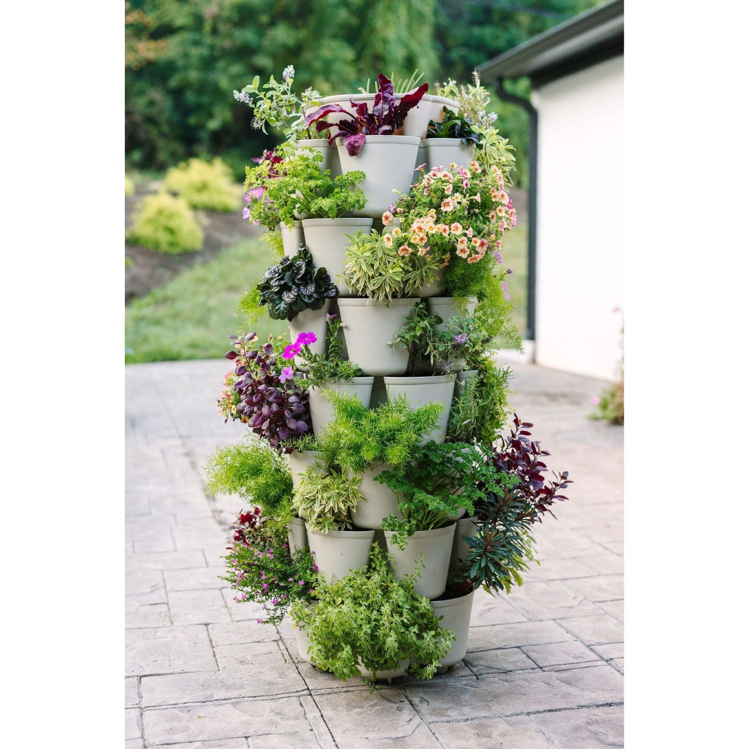 A beige 7 tier greenstalks planter with seven vertical levels filled with green and flowering plants on a bricked patio against a blurred background of greenery  - 